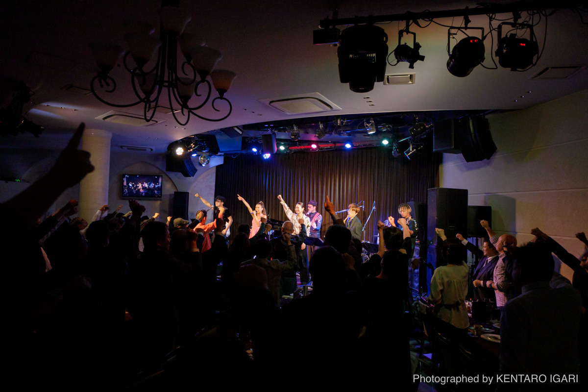 手を上げる観客とステージ全体を写したライブ風景